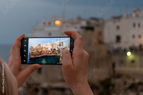 Town on the cliffs. Traveling concept background with old traditional houses, dramatic sky, Mediterranean Sea and blonde hair tourist girl taking photo with smarthphone at evening