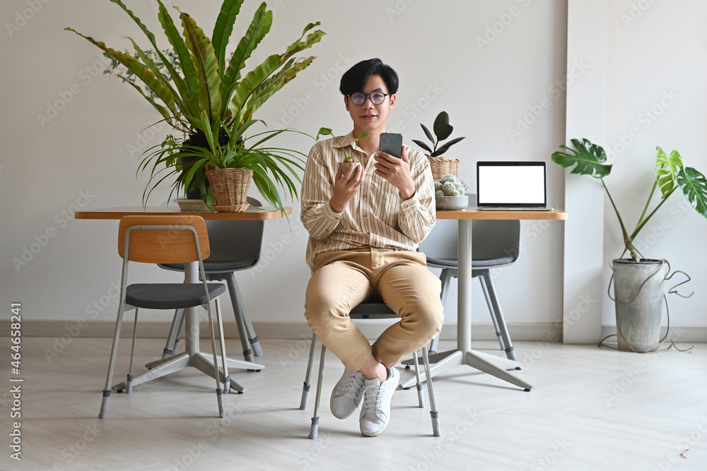 Photo of a young man showing a small plant and smartphone in hand while sitting over the working desk surrounded by a computer laptop and potted plant as a background. Photo of a young man showing a small plant and smartphone in hand while sitting over the working desk surrounded by a computer laptop and potted plant as a background.