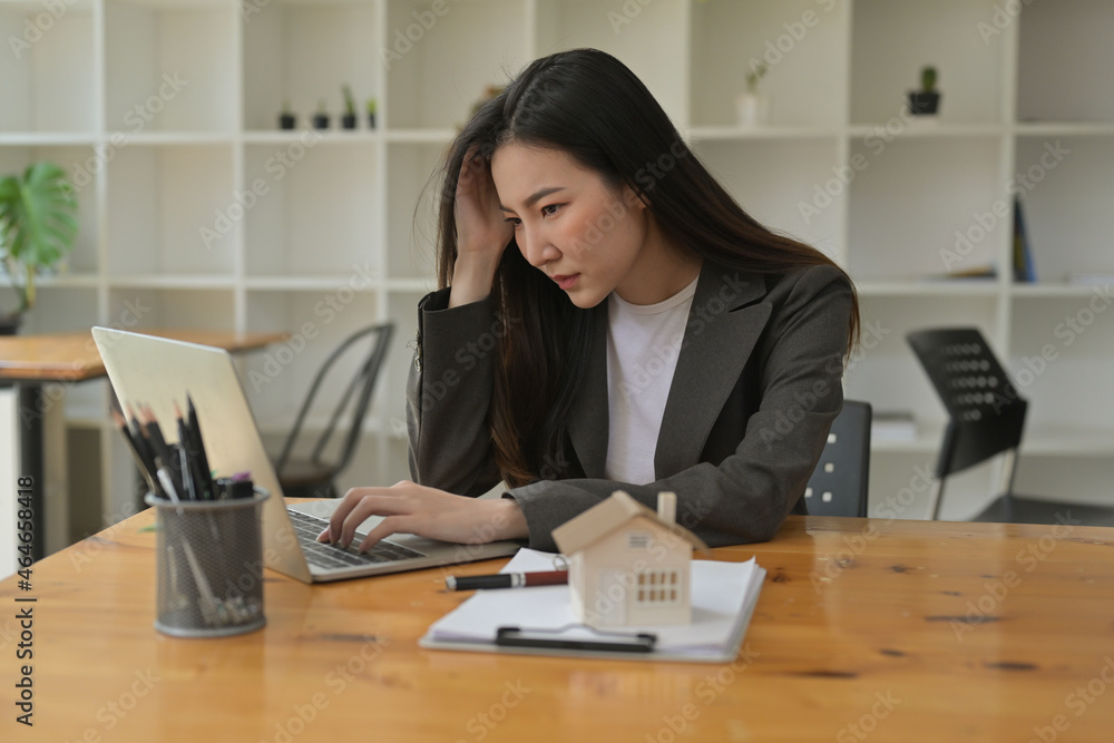 Photo of a stressed businesswoman while using a computer laptop at the wooden working desk surrounded by a clipboard, pencil holder, pen and house model. Photo of a stressed businesswoman while using a computer laptop at the wooden working desk surrounded by a clipboard, pencil holder, pen and house model.