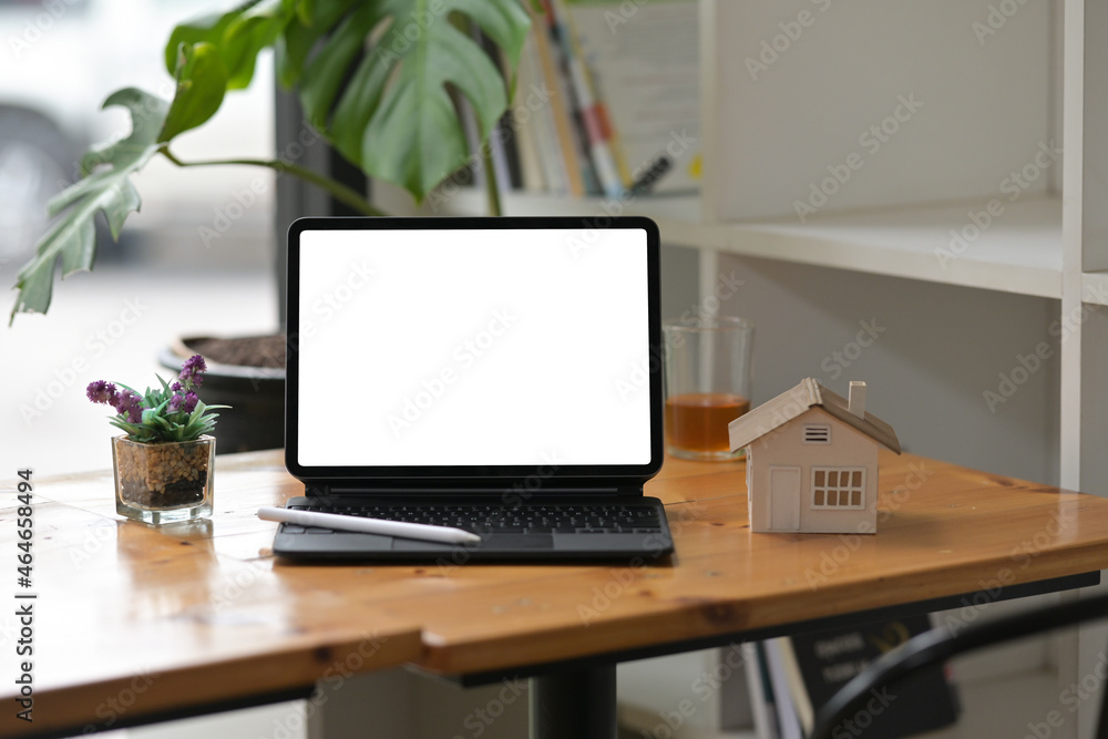 Photo of a white blank screen digital tablet with a keyboard case putting together with a stylus pen on a wooden working desk surrounded by a house model and small cup of plant. Photo of a white blank screen digital tablet with a keyboard case putting together with a stylus pen on a wooden working desk surrounded by a house model and small cup of plant.