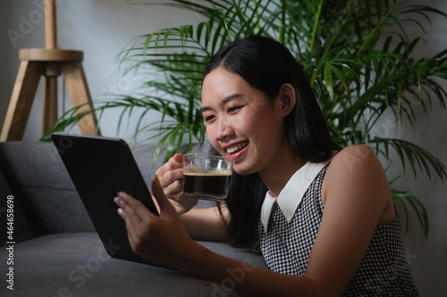 Portrait of beautiful Asian woman drinking a coffee while relaxing with a digital tablet on the cozy sofa.