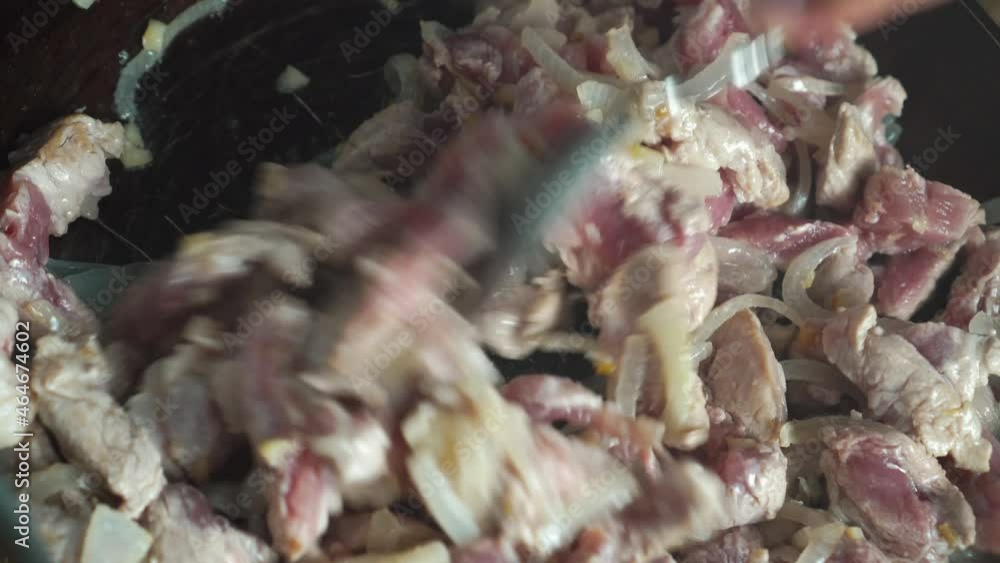 Pieces of pork flesh with chopped onions during frying in an old frying pan, top view. A woman's hand turns it over with a spoon and mixes it so that it doesn't burn. Potatoes are being cooked nearby. Pieces of pork flesh with chopped onions during frying in an old frying pan, top view. A woman's hand turns it over with a spoon and mixes it so that it doesn't burn. Potatoes are being cooked nearby.