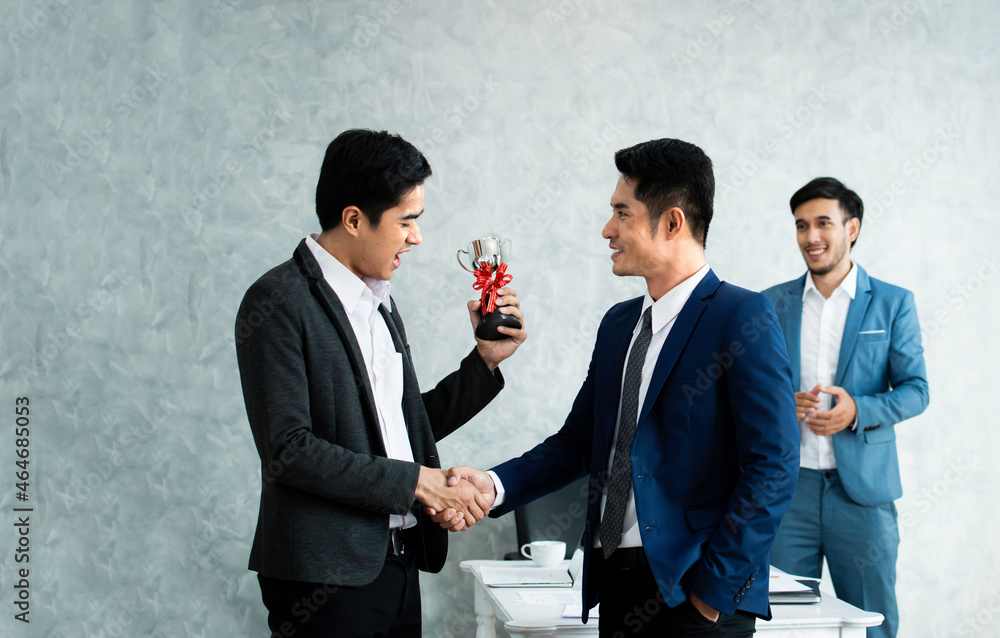 Successful business men shake hand and hold trophy while their colleagues applauding and smiling in the background. Smiling businesspeople congratulate colleague with job success or achievement. Successful business men shake hand and hold trophy while their colleagues applauding and smiling in the background. Smiling businesspeople congratulate colleague with job success or achievement.