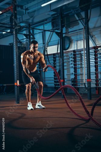 Front view of young man, muscled athlete, bodybuilder training alone at sport gym, indoors. Concept of sport, activity, healthy lifestyle