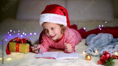 A little smiling girl in Santa's red hat and red christmas clothing lies on the bed and writes a letter to Santa Claus. New Year. hildren's Christmas. Atmosphere. Home. photo