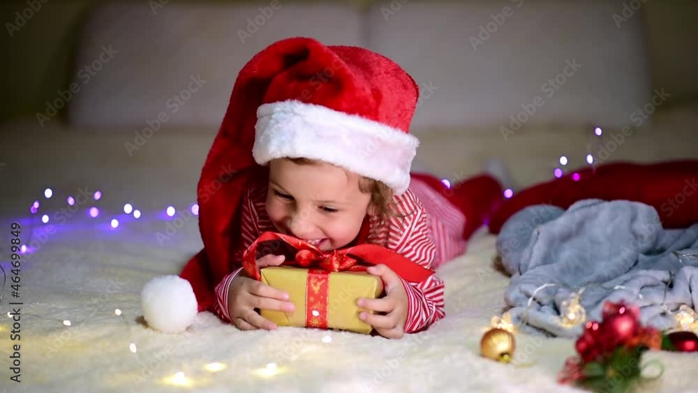 Children's Christmas. A little smiling girl in Santa's red hat and red christmas clothing lies on the bed with a gift. Preparation for the celebration. New Year. Atmosphere. Home. Children's Christmas. A little smiling girl in Santa's red hat and red christmas clothing lies on the bed with a gift. Preparation for the celebration. New Year. Atmosphere. Home.