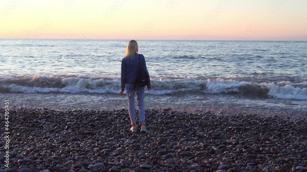 girl stands by sea and dreams. Romantic walk along beautiful shore. Strong waves on sea. A beautiful slender blonde in denim jacket with leather backpack stands on shore and looks at sunset. girl stands by sea and dreams. Romantic walk along beautiful shore. Strong waves on sea. A beautiful slender blonde in denim jacket with leather backpack stands on shore and looks at sunset.