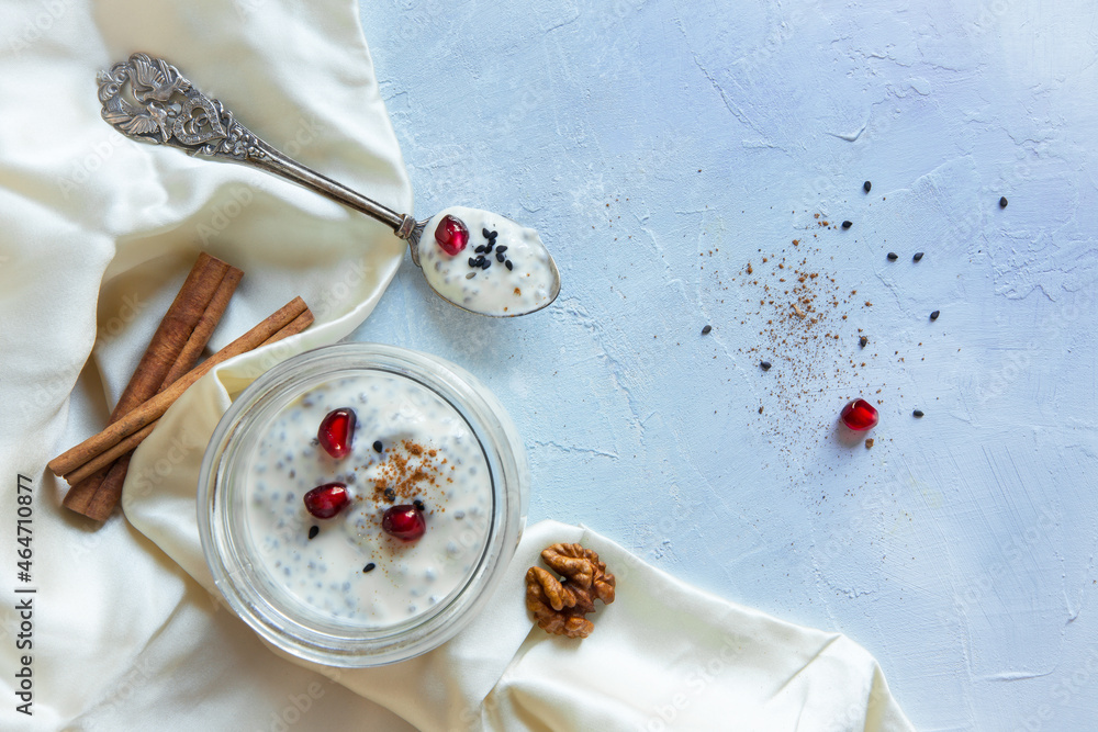 Flat lay style high key composition of diary free vegan soy yogurt with chia seeds, cinnamon and pomegranate seeds with silk cloth and silver spoon on a light painted background