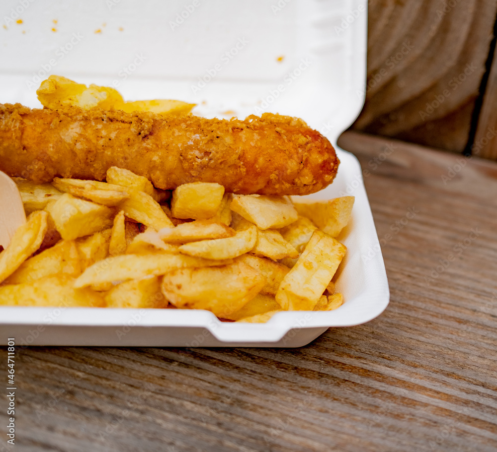 Close and selective focus on a polystyrene carton comprising a battered jumbo sausage and traditional chip shop chips cooked in beef dripping on an outdoor wooden table Close and selective focus on a polystyrene carton comprising a battered jumbo sausage and traditional chip shop chips cooked in beef dripping on an outdoor wooden table