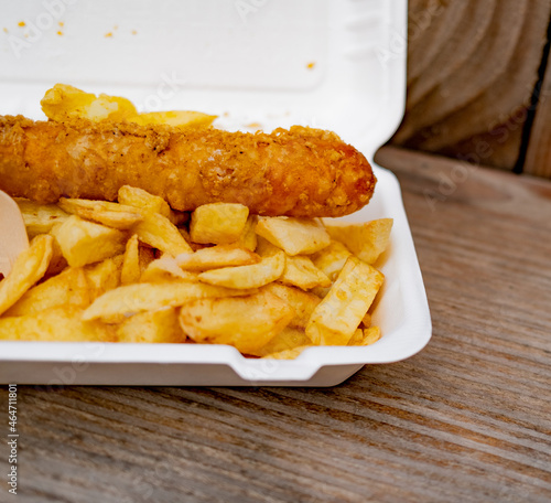 Close and selective focus on a polystyrene carton comprising a battered jumbo sausage and traditional chip shop chips cooked in beef dripping on an outdoor wooden table