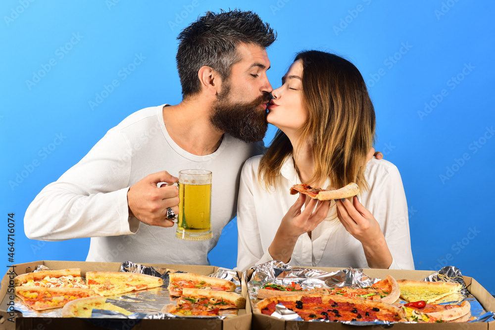 Loving couple sitting at table kissing and eating pizza. Pretty young girl in white shirt holds slice yummy pizza and handsome boyfriend in white pullover with glass of beer. Boxes with pizza on table Loving couple sitting at table kissing and eating pizza. Pretty young girl in white shirt holds slice yummy pizza and handsome boyfriend in white pullover with glass of beer. Boxes with pizza on table