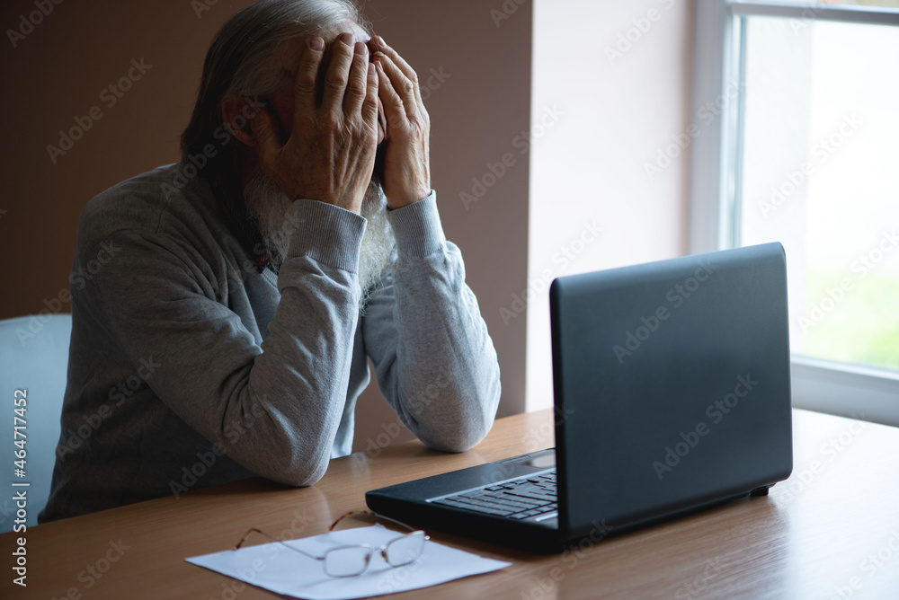 A tired, broken, resigned, preoccupied elderly man sits in front of a laptop, computer and covers his face with his hand. The sad senior checks his account balance. A tired, broken, resigned, preoccupied elderly man sits in front of a laptop, computer and covers his face with his hand. The sad senior checks his account balance.