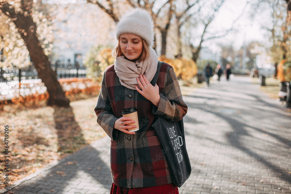 A young girl in a white hat and a plaid shirt coat walks along an autumn alley in the city with a shopper and enjoys a sunny day. He holds a glass of coffee in his hands