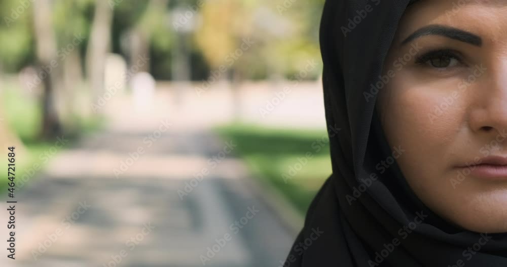 Half face portrait of muslim woman looking at camera, close-up. Arab woman pose in black hijab at city street. Serious, sad saudi female in traditional headscarf veil outdoors. With copy space text Half face portrait of muslim woman looking at camera, close-up. Arab woman pose in black hijab at city street. Serious, sad saudi female in traditional headscarf veil outdoors. With copy space text