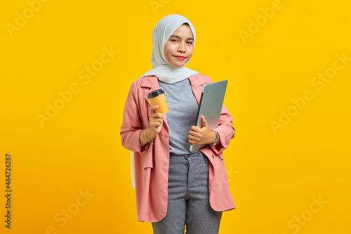 Portrait of smilling young Asian woman holding laptop and coffee isolated over yellow background