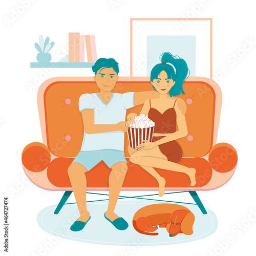 A guy with a girl in comfortable home clothes is sitting on a sofa and eating mouth-watering popcorn from a striped box, and an orange dachshund dog sleeps at their feet. Weekend home holidays.