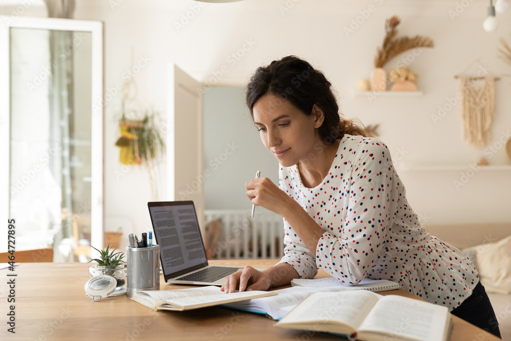 Focused adult female student doing homework, reading books at laptop, taking notes. New mother learning online from home, getting new occupation, preparing for college test. Education concept Focused adult female student doing homework, reading books at laptop, taking notes. New mother learning online from home, getting new occupation, preparing for college test. Education concept