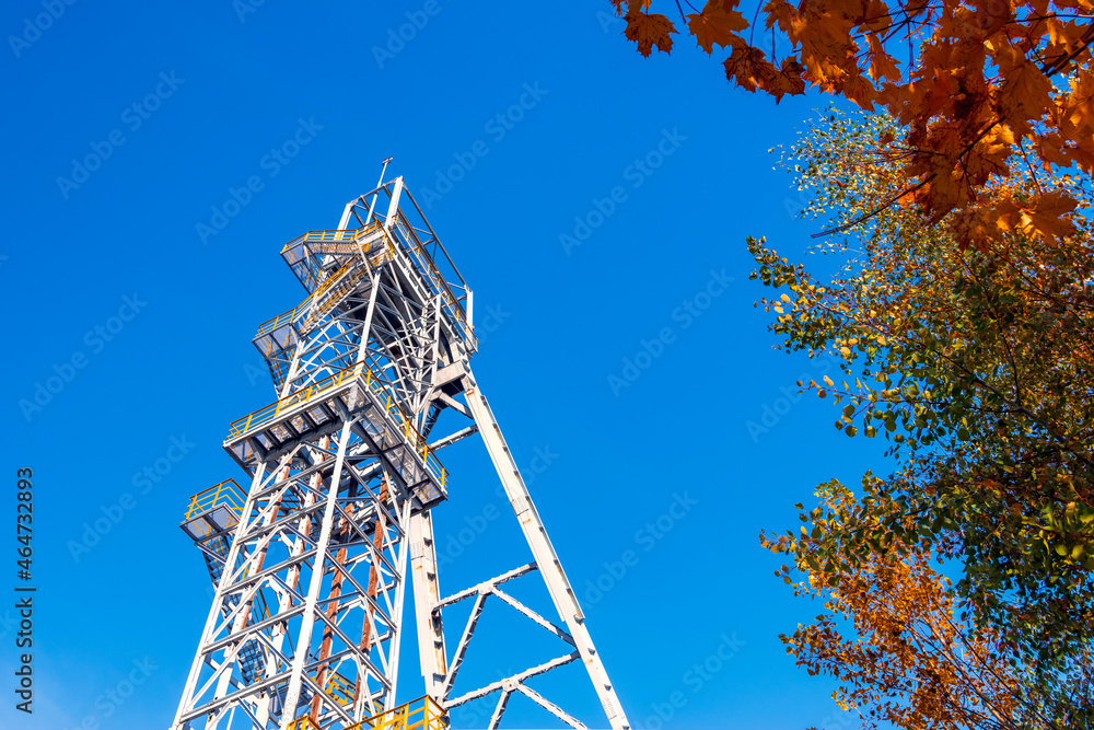 Mine shaft tower 'Krystyn' in former coal mine 'Michal' in Siemianowice, Silesia, Poland against blue sky. Beautiful, autumn tree in the foreground.