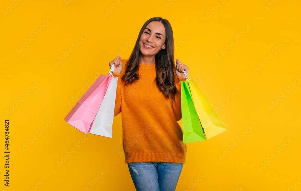 Cute fashionable young smiling woman holds many colorful shopping paper bags in hand while she is isolated on yellow background
