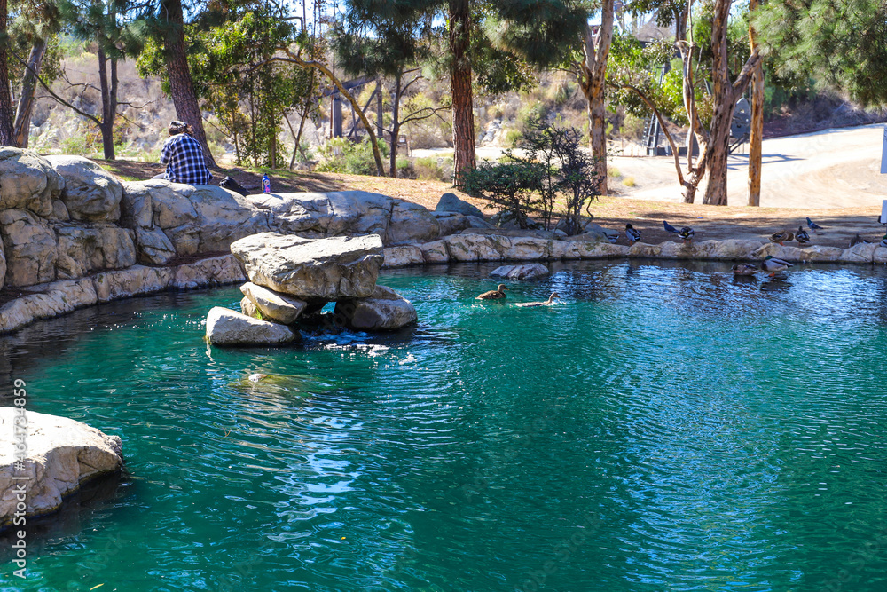a circular stone lagoon filled with gorgeous blue water surrounded by lush green and autumn colored trees with a woman sitting on the edge at Kenneth Hahn Recreation Area in Los Angeles CA