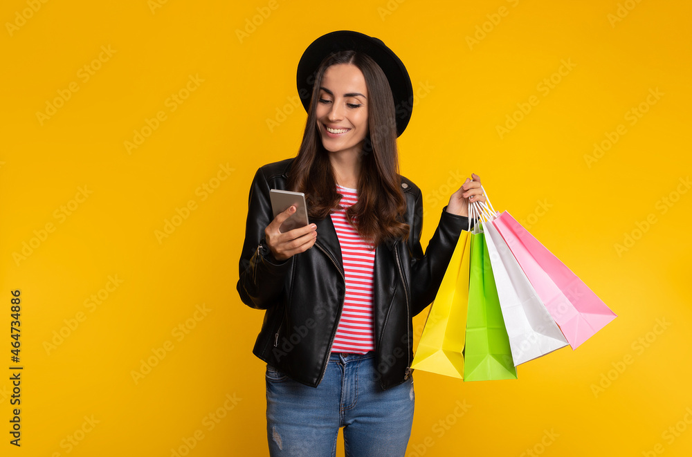 Trendy beautiful young hipster girl in black hat with paper bags in hand is using her smart phone for online shopping while isolated on yellow background