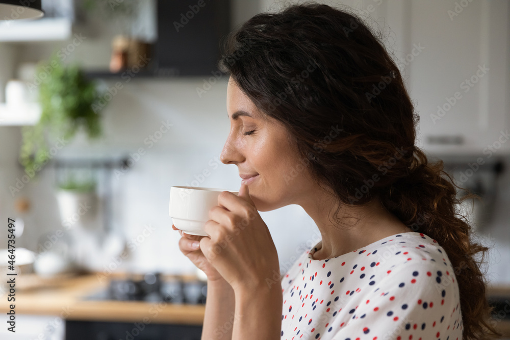 Happy peaceful woman enjoying morning coffee in kitchen, holding cup, inhaling aroma with closed eyes. Homeowner relaxing over cup of hot beverage at home, smiling, thinking and dreaming Happy peaceful woman enjoying morning coffee in kitchen, holding cup, inhaling aroma with closed eyes. Homeowner relaxing over cup of hot beverage at home, smiling, thinking and dreaming