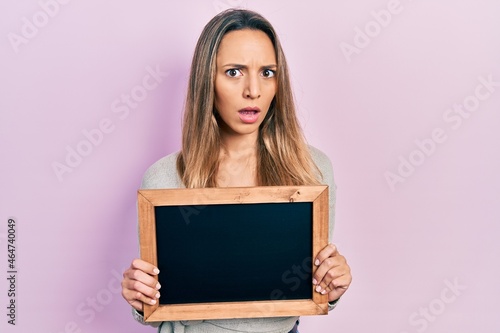 Beautiful hispanic woman holding small blackboard in shock face, looking skeptical and sarcastic, surprised with open mouth