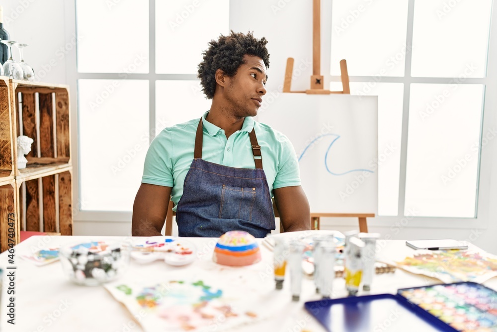 Young african american man sitting on the table at art studio looking to side, relax profile pose with natural face with confident smile. Young african american man sitting on the table at art studio looking to side, relax profile pose with natural face with confident smile.
