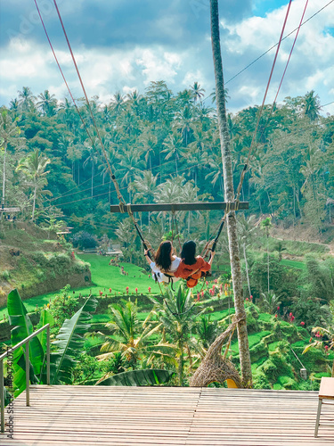 Vacation concept. Happy two young womans in dress swinging at palm with bali rice terraces background, Bali, Indonesia.