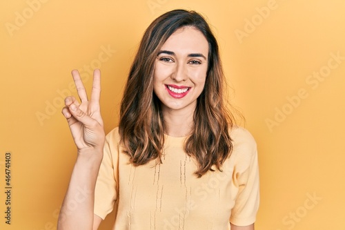 Young hispanic girl wearing casual clothes smiling with happy face winking at the camera doing victory sign. number two.