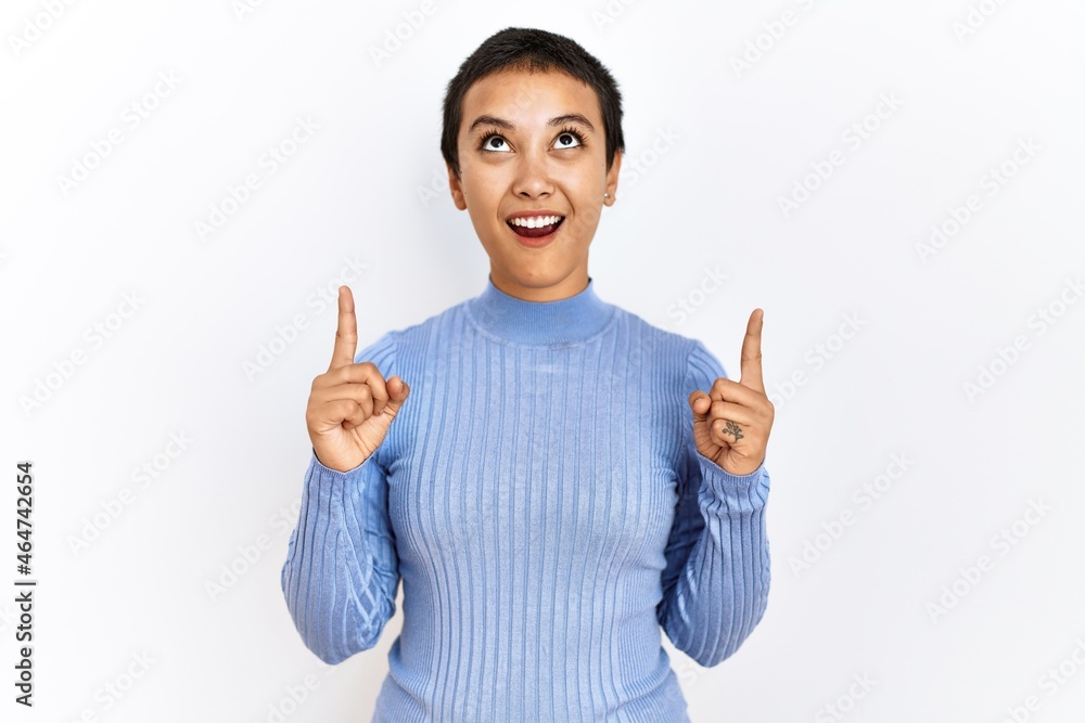Young hispanic woman with short hair standing over isolated background amazed and surprised looking up and pointing with fingers and raised arms. Young hispanic woman with short hair standing over isolated background amazed and surprised looking up and pointing with fingers and raised arms.