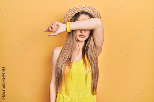 Young blonde girl wearing summer hat covering eyes with arm, looking serious and sad. sightless, hiding and rejection concept