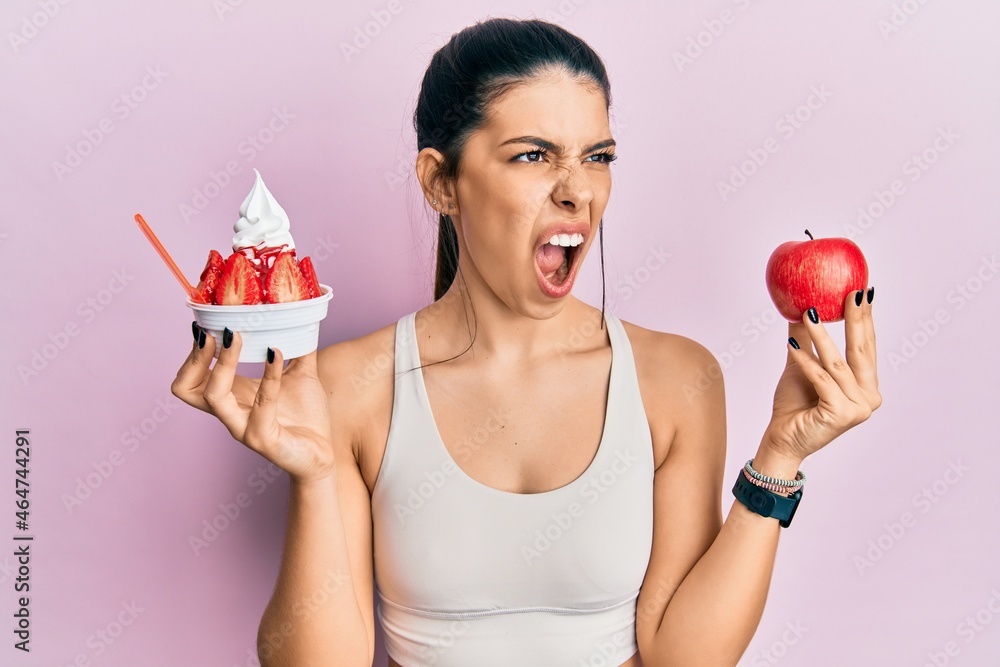 Young hispanic woman wearing sportswear holding apple and ice cream angry and mad screaming frustrated and furious, shouting with anger. rage and aggressive concept. Young hispanic woman wearing sportswear holding apple and ice cream angry and mad screaming frustrated and furious, shouting with anger. rage and aggressive concept.