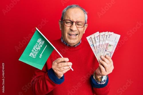 Handsome senior man with grey hair holding saudi arabia flag and riyal banknotes winking looking at the camera with sexy expression, cheerful and happy face.
