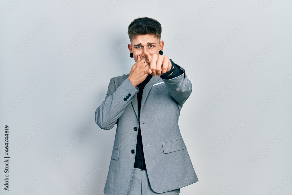 Young caucasian boy with ears dilation wearing business jacket laughing at you, pointing finger to the camera with hand over mouth, shame expression Young caucasian boy with ears dilation wearing business jacket laughing at you, pointing finger to the camera with hand over mouth, shame expression