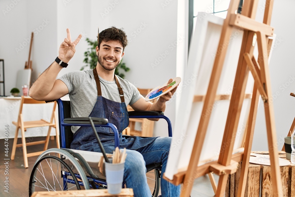 Young hispanic man sitting on wheelchair painting at art studio smiling with happy face winking at the camera doing victory sign with fingers. number two.