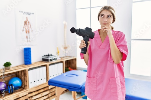 Young physiotherapist woman holding therapy massage gun at wellness center serious face thinking about question with hand on chin  thoughtful about confusing idea