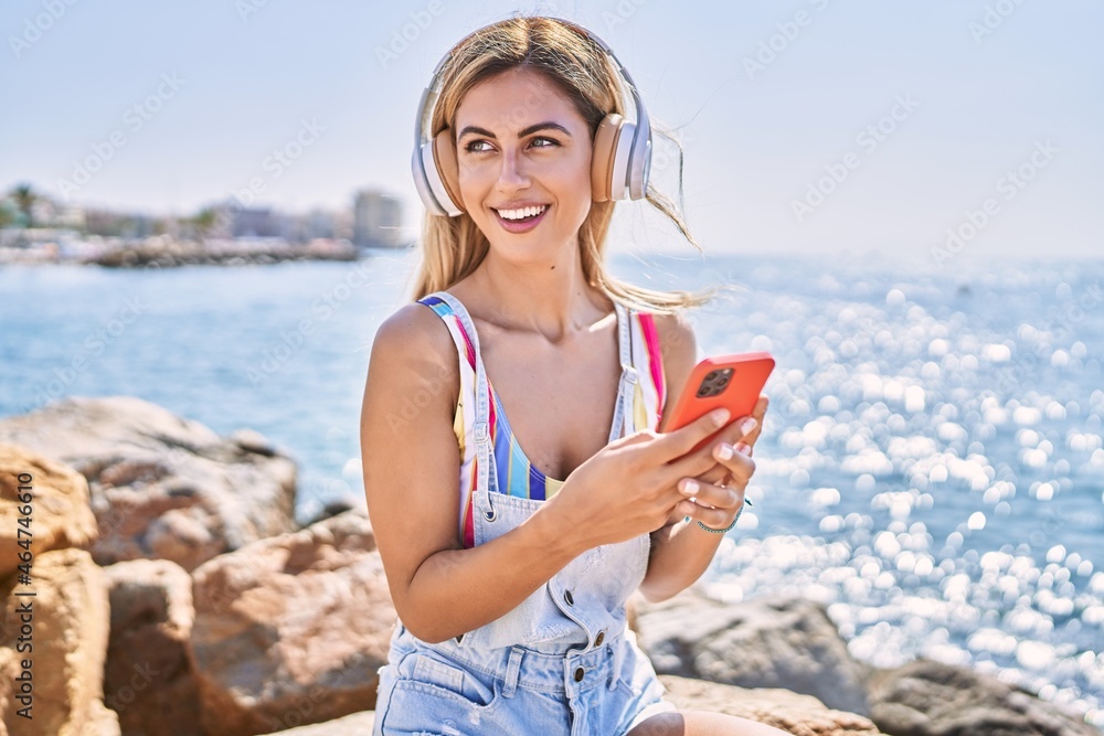 Young blonde girl listening to music sitting on the rock at the beach.