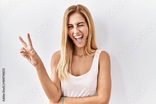Blonde beautiful young woman standing over white isolated background smiling with happy face winking at the camera doing victory sign. number two.