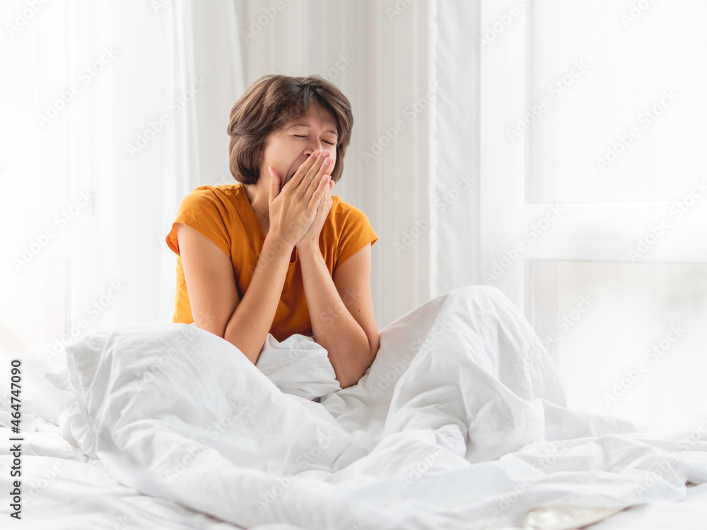 Sleepy woman is yawning in bed, covered with white blanket. It's hard to wake up early in morning.