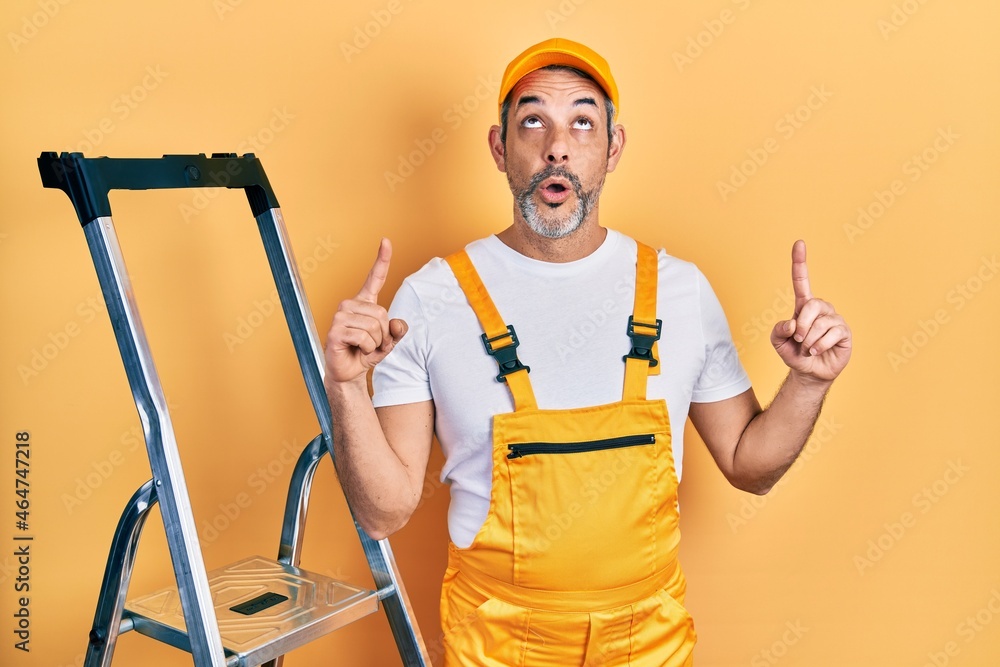 Handsome middle age man with grey hair holding ladder amazed and surprised looking up and pointing with fingers and raised arms. Handsome middle age man with grey hair holding ladder amazed and surprised looking up and pointing with fingers and raised arms.