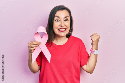 Middle age hispanic woman holding pink cancer ribbon screaming proud, celebrating victory and success very excited with raised arms