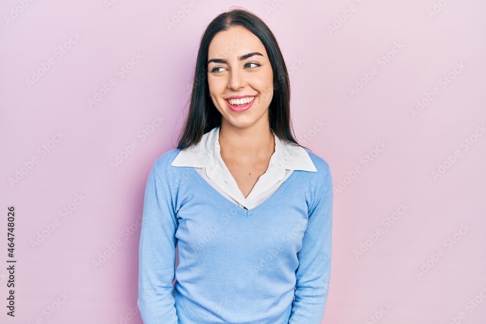 Beautiful woman with blue eyes standing over pink background looking away to side with smile on face, natural expression. laughing confident.