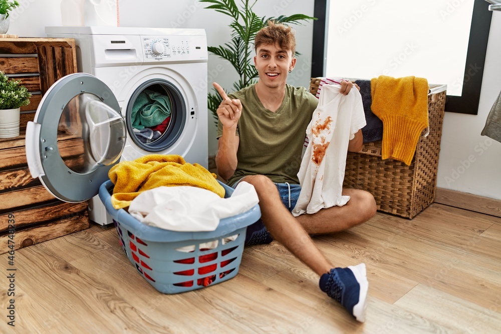 Young caucasian man doing laundry holding t shirt with stain smiling happy pointing with hand and finger to the side Young caucasian man doing laundry holding t shirt with stain smiling happy pointing with hand and finger to the side