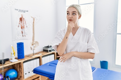 Young caucasian woman working at pain recovery clinic looking confident at the camera smiling with crossed arms and hand raised on chin. thinking positive.