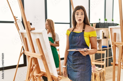 Young hispanic woman at art classroom scared and amazed with open mouth for surprise, disbelief face