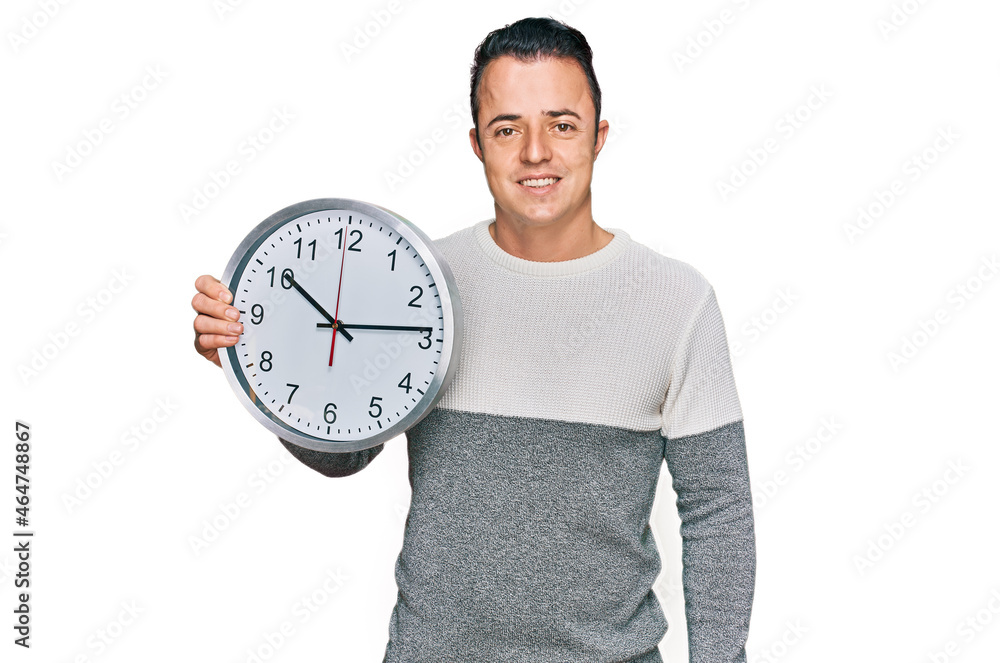 Handsome young man holding big clock looking positive and happy standing and smiling with a confident smile showing teeth Handsome young man holding big clock looking positive and happy standing and smiling with a confident smile showing teeth
