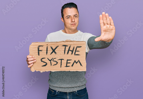 Handsome young man holding fix the system banner cardboard with open hand doing stop sign with serious and confident expression, defense gesture photo