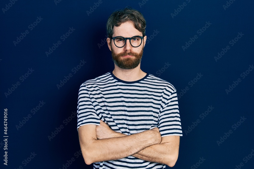 Caucasian man with beard wearing striped t shirt and glasses skeptic and nervous, disapproving expression on face with crossed arms. negative person. Caucasian man with beard wearing striped t shirt and glasses skeptic and nervous, disapproving expression on face with crossed arms. negative person.