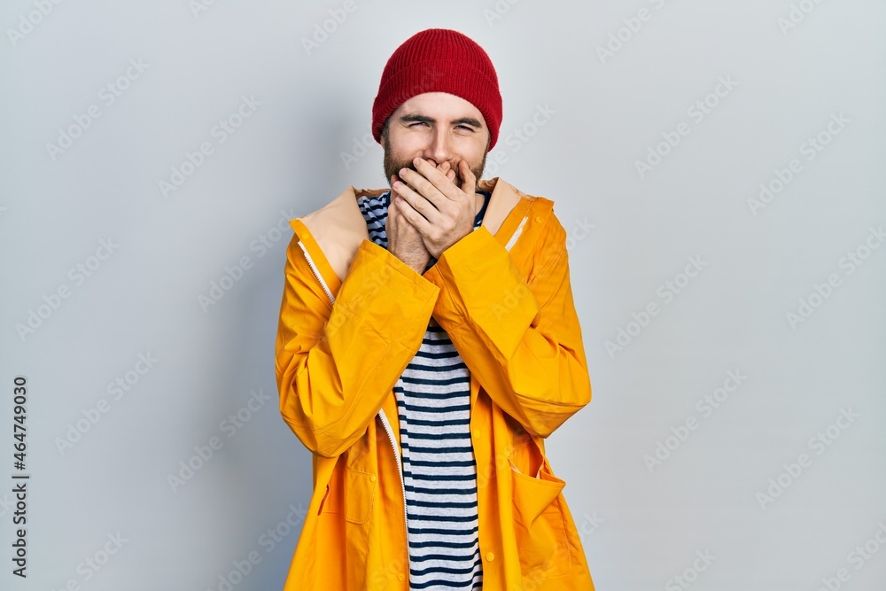 Caucasian man with beard wearing yellow raincoat laughing and embarrassed giggle covering mouth with hands, gossip and scandal concept